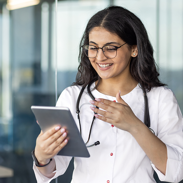 Smiling female doctor using tablet for video call in modern office setting wearing glasses and medical coat
